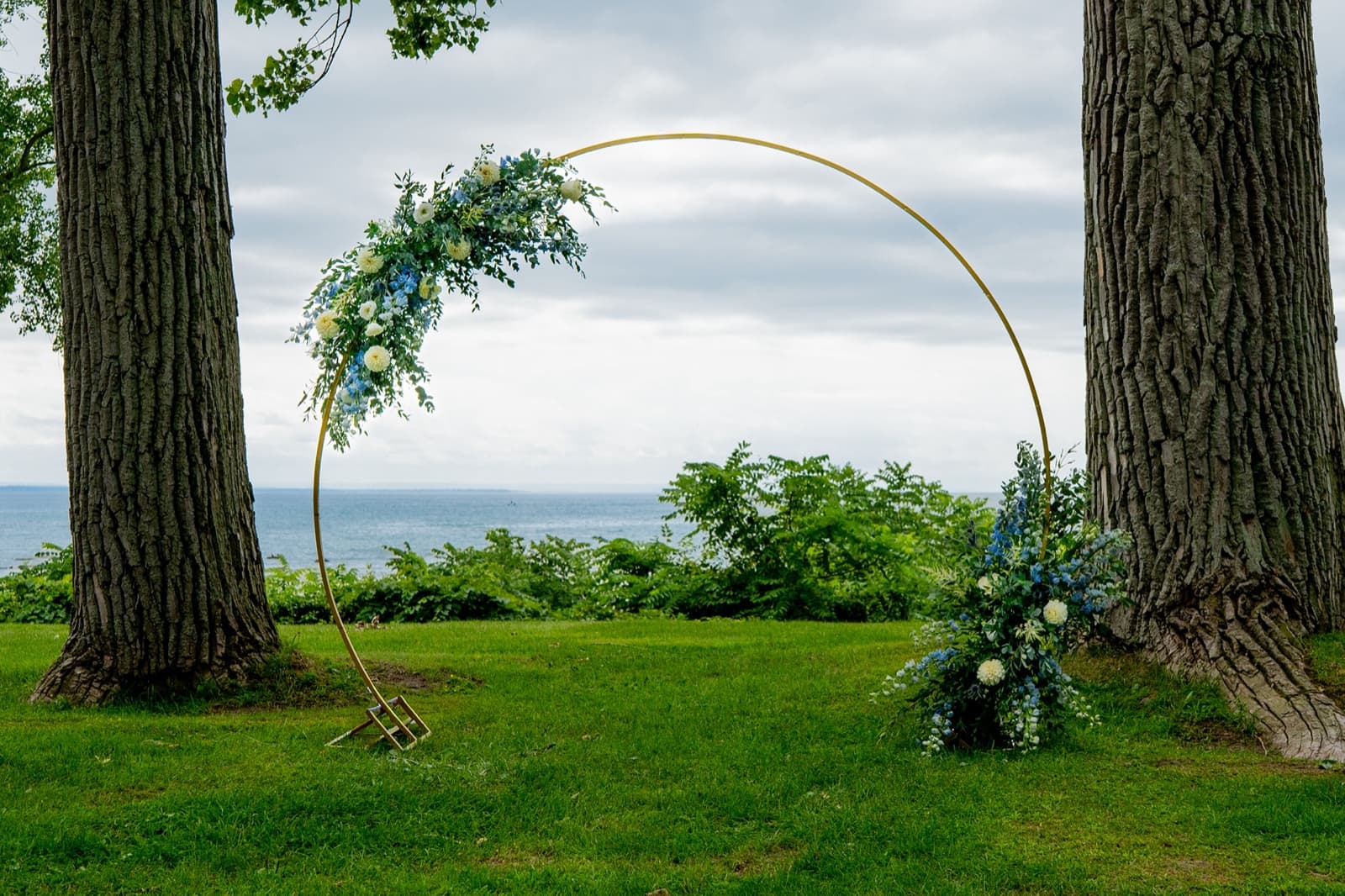 Gold circular wedding arch dressed with blue and white florals on a lakeside lawn
